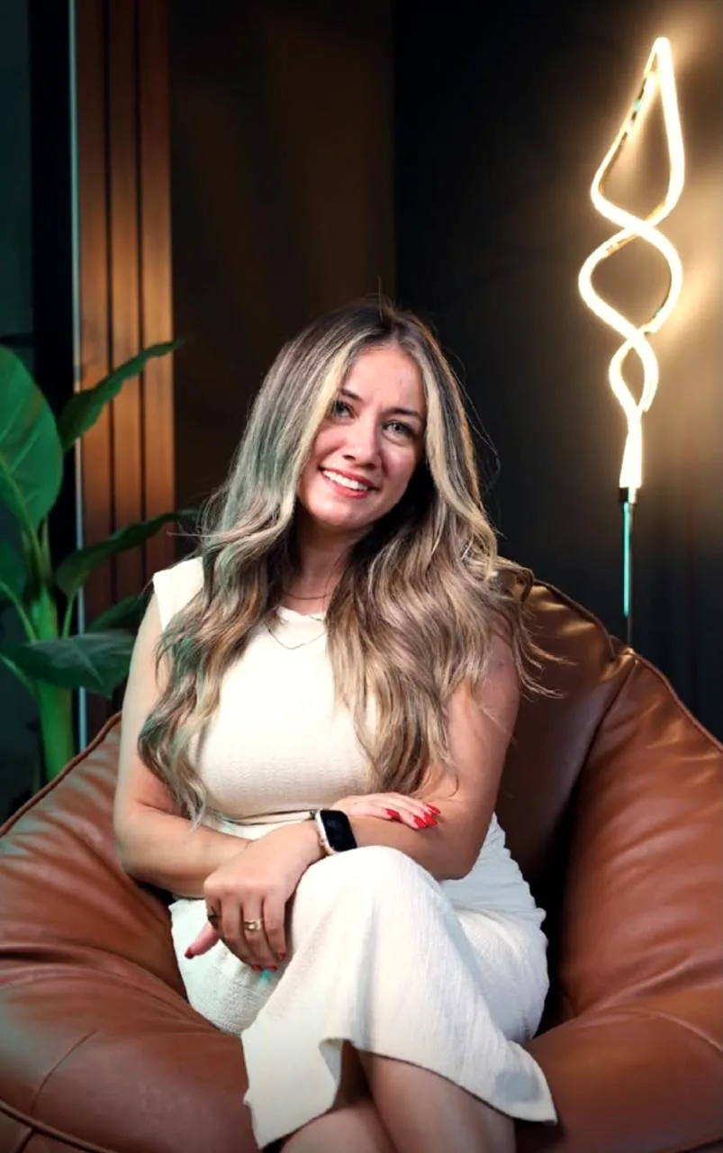 Dr. Maggy Maher seated in a leather armchair, smiling toward the camera, with a spiral neon lamp behind her.